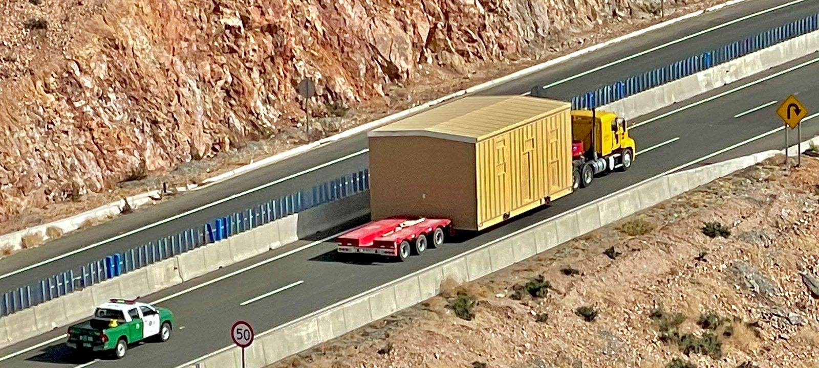 Aerial view of a Logística Inversol convoy moving an oversized module along a mountain highway, with a Carabineros (Chile's national police) patrol escorting in the opposite lane